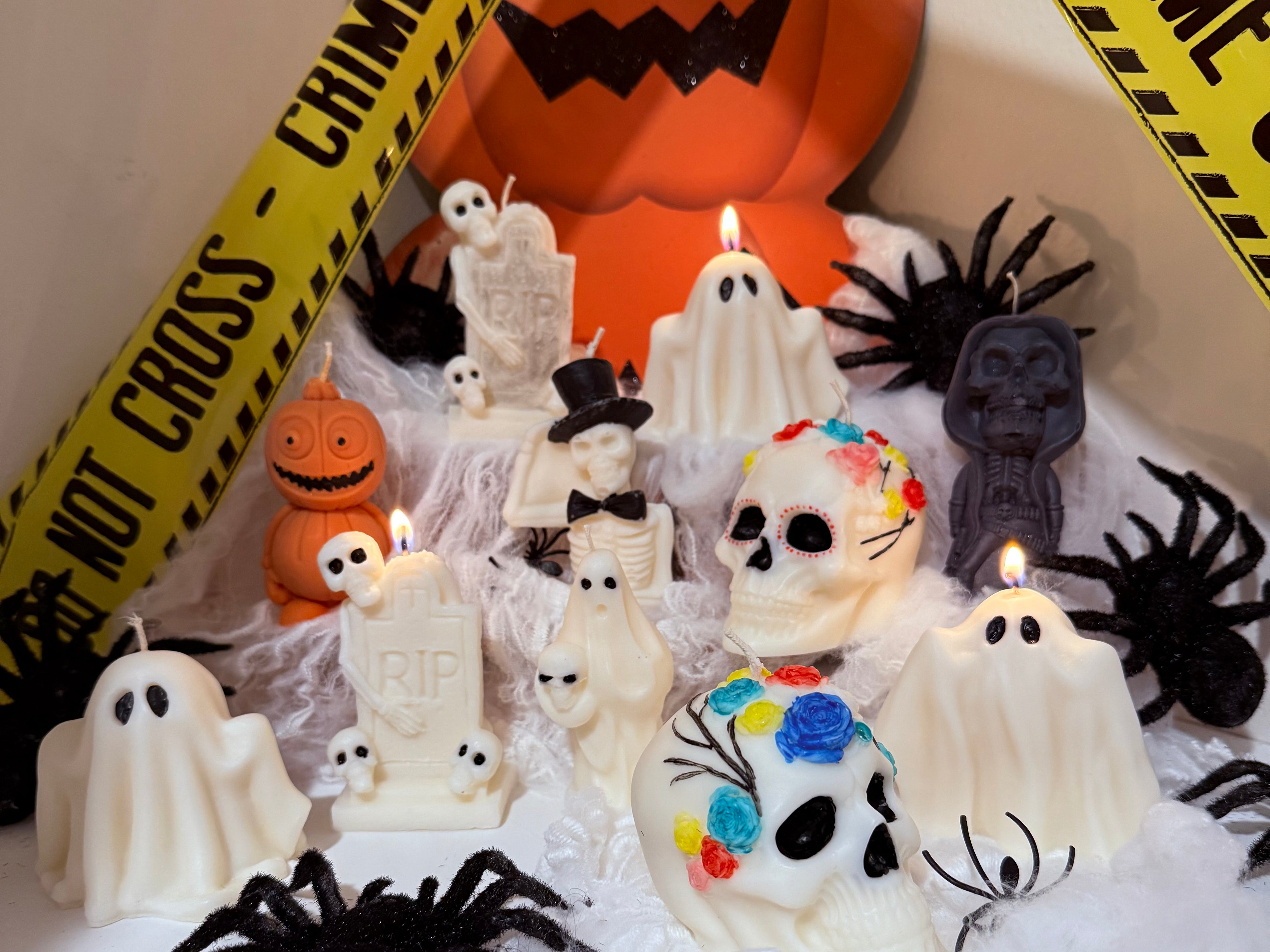 A collection of Halloween-themed candles arranged in a spooky display with white ghost candles, skull candles decorated with colorful floral details, tombstone candles, black spider decorations, and a large orange jack-o'-lantern candle in the background. Yellow caution tape props frame the scene.